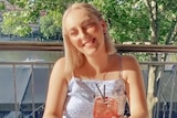 A smiling young blonde woman with a drink in her hand sits in the sun.