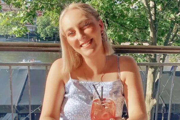 A young blonde woman sits in the sun with a drink in her hand, smiling.