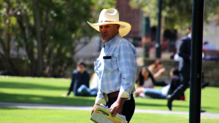 Donald, wearing a checked shirt, large white cowboy hat and over-sized belt buckle, walks with grass in the background.