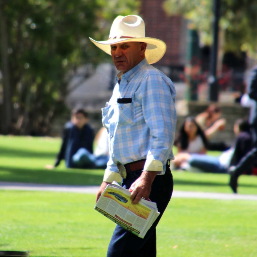 Donald, wearing a checked shirt, large white cowboy hat and over-sized belt buckle, walks with grass in the background.