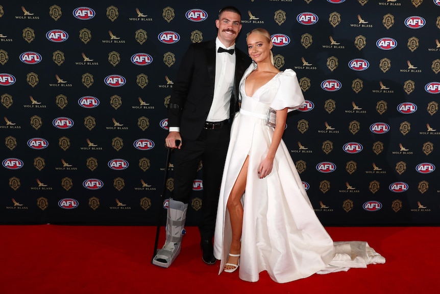 A Gold Coast AFL player in a tuxedo and a moon boot, holding a walking stick next to his partner in a white dress.