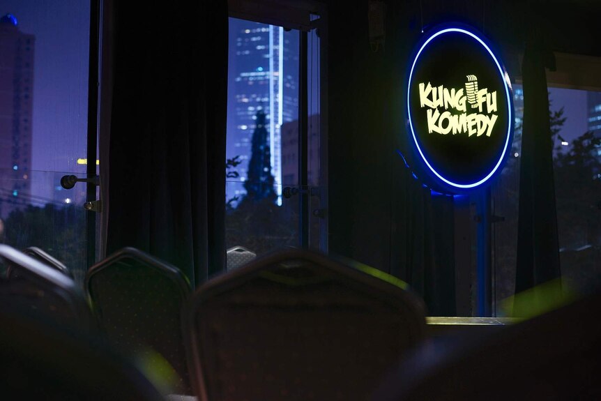 Seats surround a small stage, sign reads 'Kung Fu Comedy', night-time city scape through windows with lit-up skyscrapers