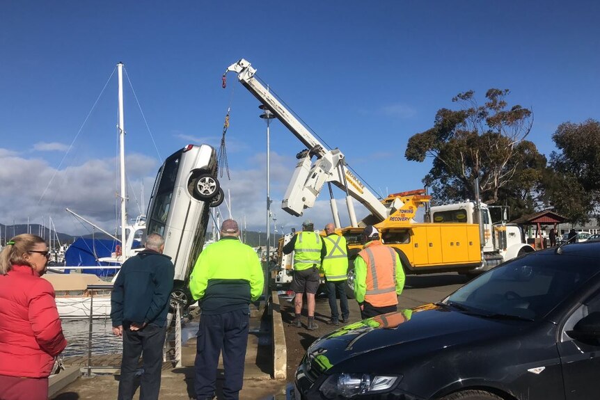 A car is lifted by crane out of marina water as people watch.