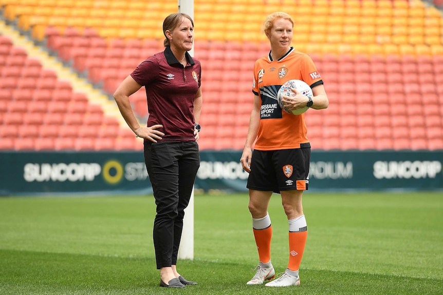 Mel Andreatta and Clare Polkinghorne stand on grass inside a stadium with yellow and red seats in the background