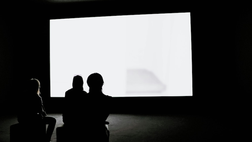 Silhouetted figures sit in a dark room in front of a big bright white screen. 