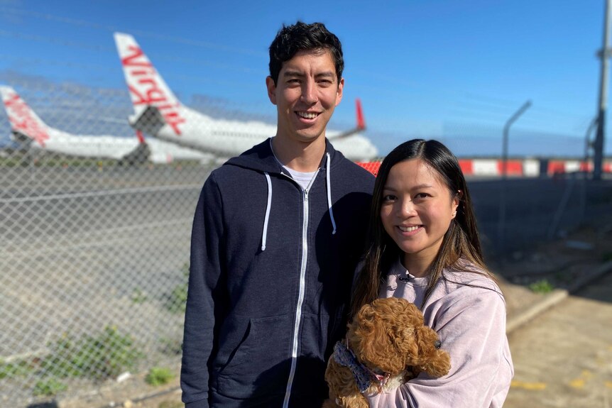 A woman holding a brown curly-haired puppy with a man behind in front of an airport fence with planes behind it