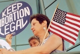 Slightly grainy photo of woman with short hair holding American flag, looking towards banner with words, 'Keep Abortion Legal'.
