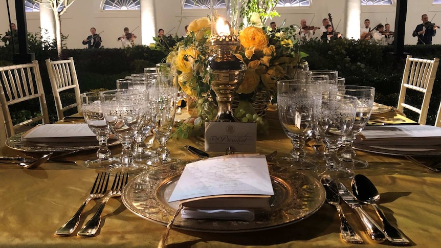 An elegant table setting featuring yellow roses and Australian wattle. Violinists play along the wall behind the table.