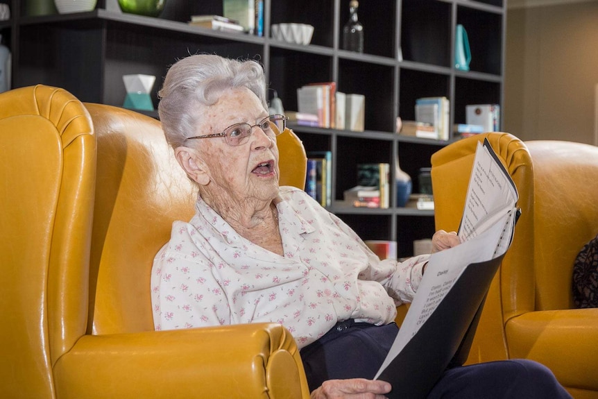 Good generic of an old woman sitting in a big padded nursing home chair singing from a plastic sleeve folder of songs