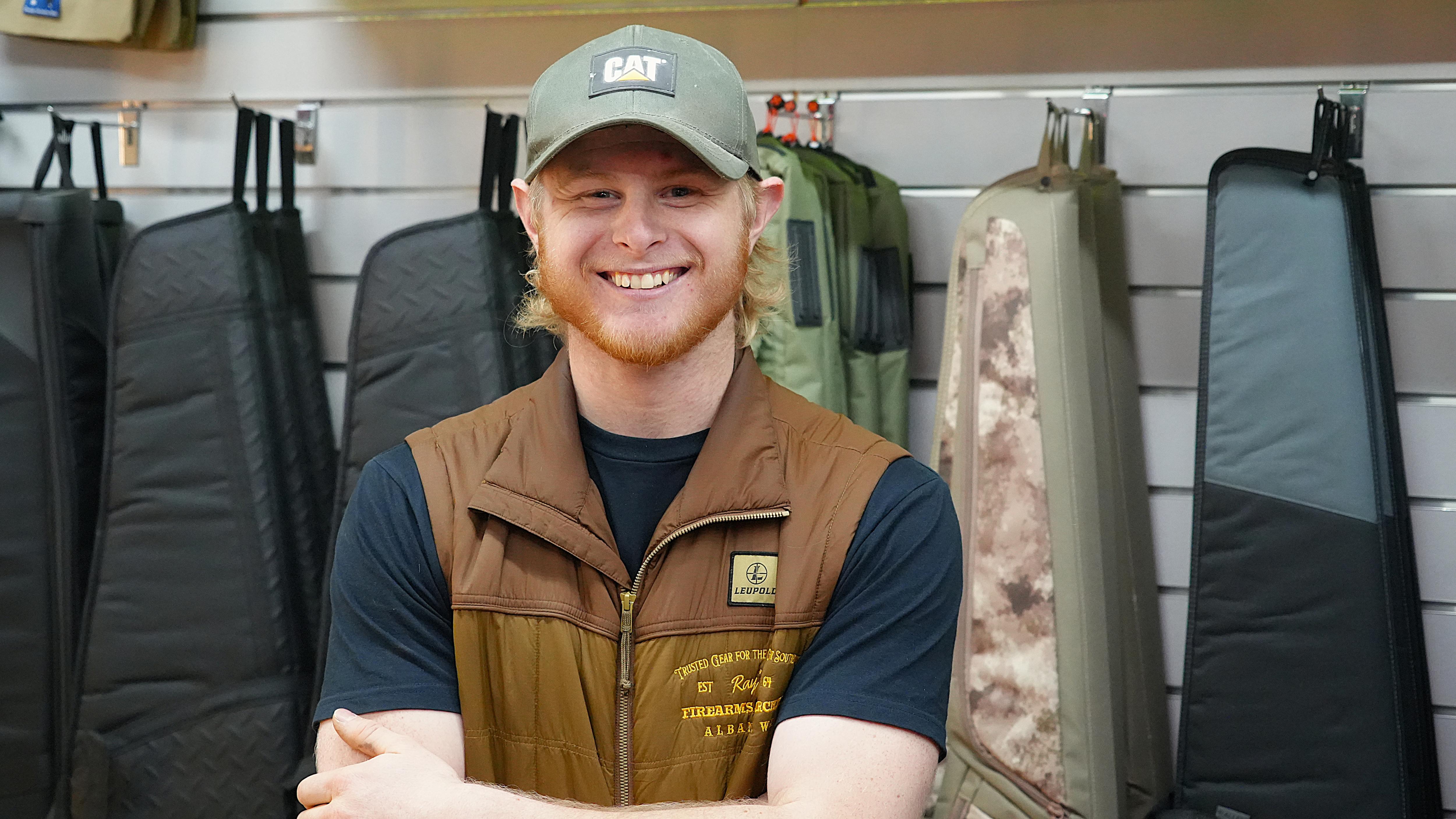 Man with short beard and blonde hair, wearing green hat and brown vest looking at camera.