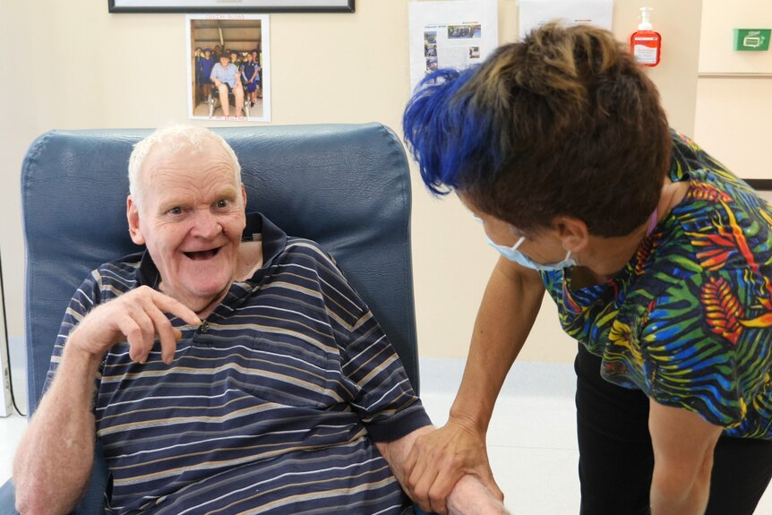 An older man sits in a chair with a big grin on his face while a woman with dyed hair attends to him.