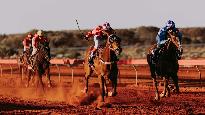Jockeys riding horses on a red dirt track at country races. 
