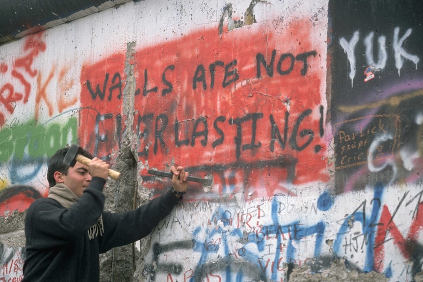 A man chisels a graffiti-covered section of the Berlin Wall that says 'Walls Are Not Everlasting!'