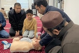 A man demonstrates CPR on a plastic dummy to a group of men in a room