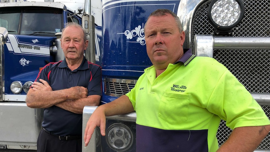 Two men with serious expressions of on their faces stand in front of two blue transport trucks