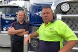 Two men with serious expressions of on their faces stand in front of two blue transport trucks