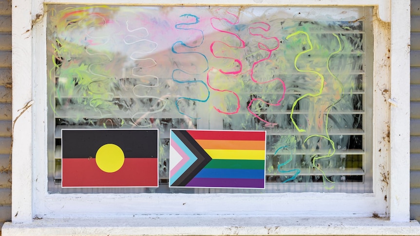 An Aboriginal and a pride flag are stuck on a window with colourful squiggles drawn on the glass around them.