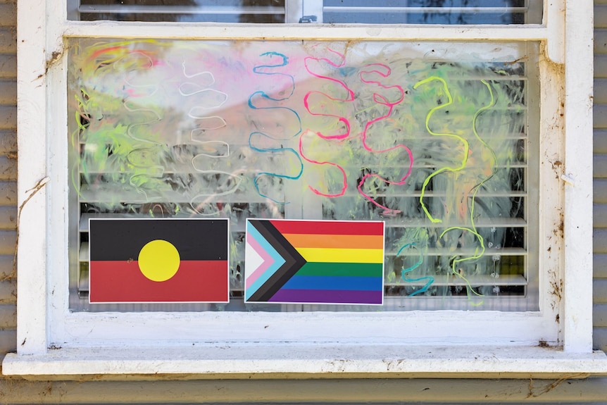An Aboriginal and a pride flag are stuck on a window with colourful squiggles drawn on the glass around them.