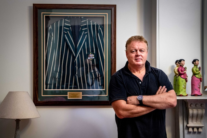 A man stands in front of a framed jacket hanging on the wall, with his arms crossed.