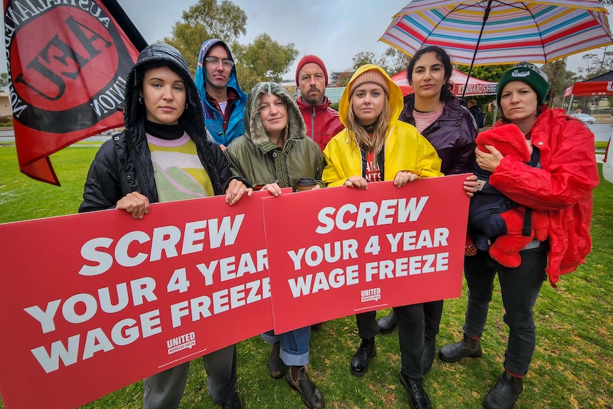 Seven people huddled together holding signs reading 'Screw your 4 year wage freeze'.