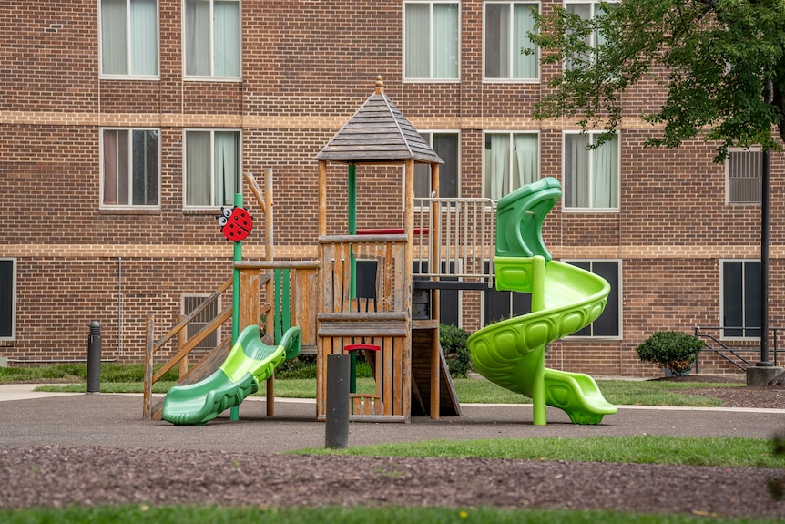Wooden playing equipment, with two green fibreglass slippery dips, in front of a large brown brick apartment building.