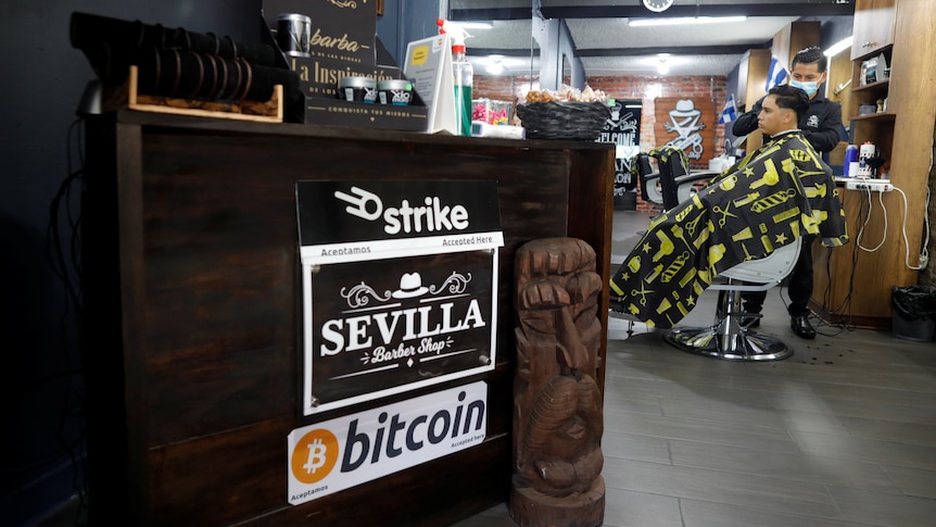 A barber in El Salvador with a man getting his haircut in the background; in the foreground a sign saying they accept bitcoin. 