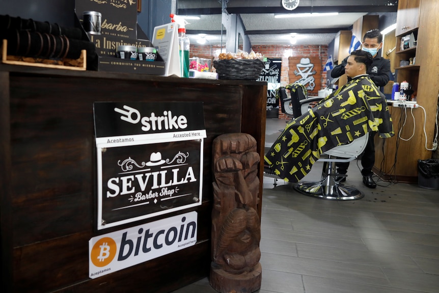 A barber in El Salvador with a man getting his haircut in the background; in the foreground a sign saying they accept bitcoin. 