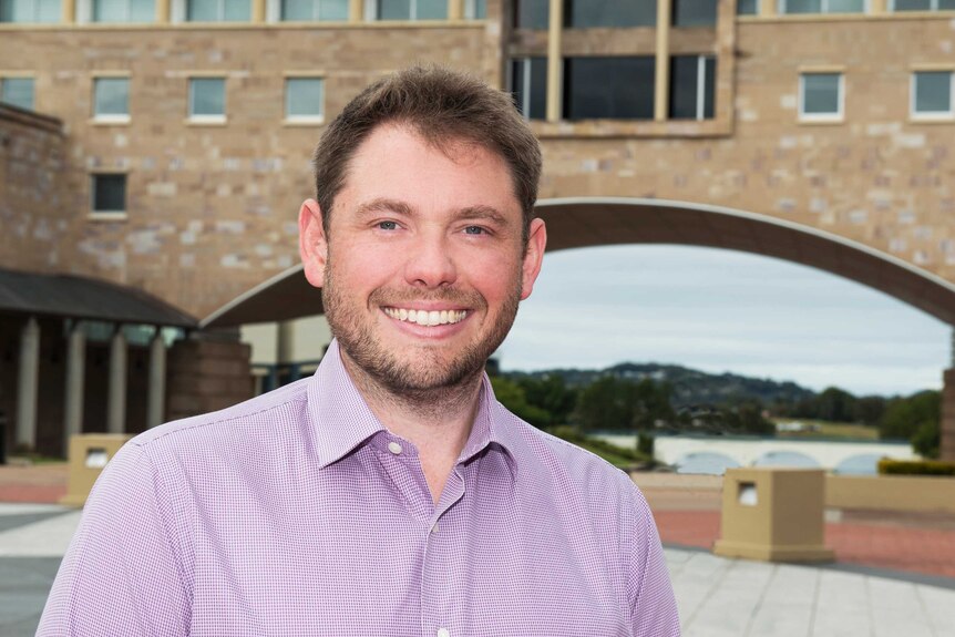 A smiling man wearing a purple checked shirt stands in a stone courtyard.