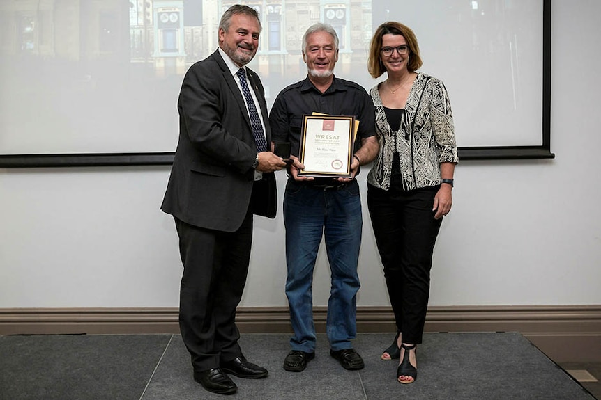 Two men and a woman stand in front of a projection screen holding a certificate and smiling.