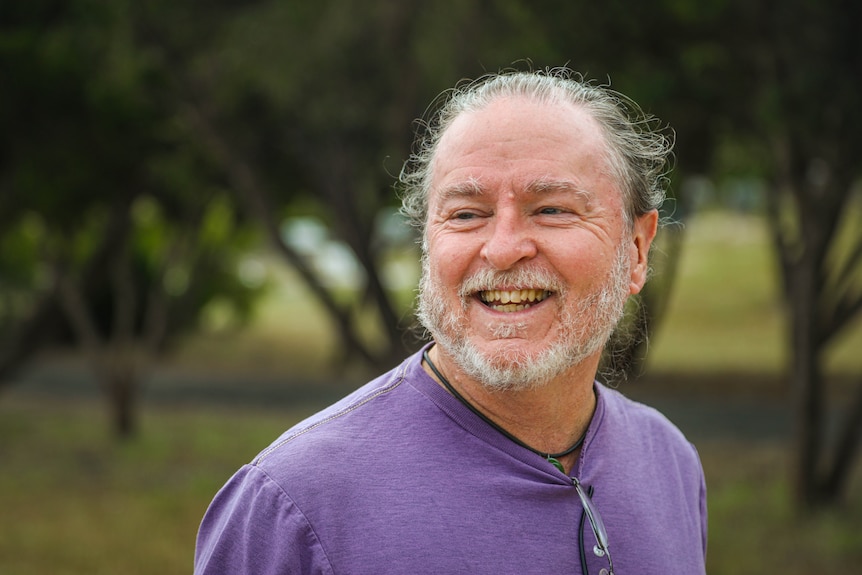 A middle aged man wearing a purple shirt smiles away from the camera.