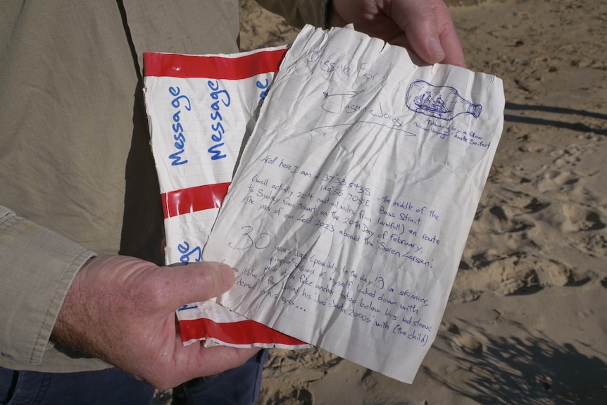 Close up of man's hands holding a crumpled handwritten letter with a drawing of a ship in a bottle.