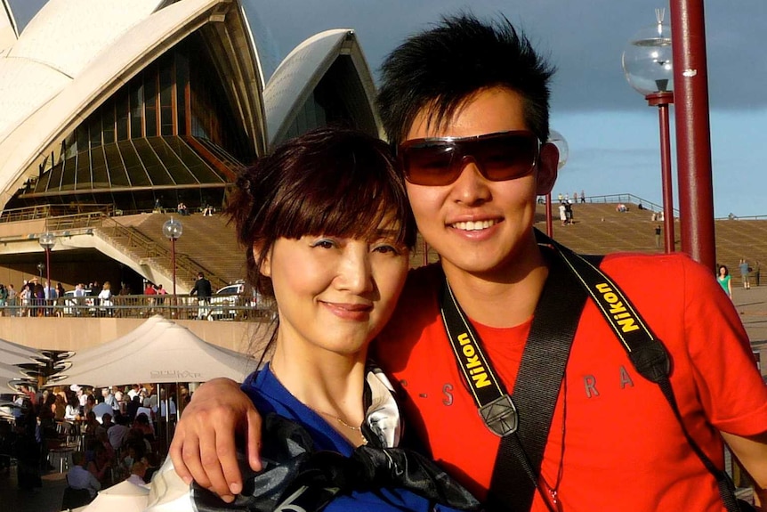 Women wearing a blue top puts arm around young man holding a camera in front of the Sydney Opera House.