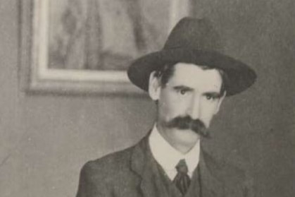 Black and white grainy photo of Henry Lawson with thick mustache and round cap, and jacket, vest and tie, standing.
