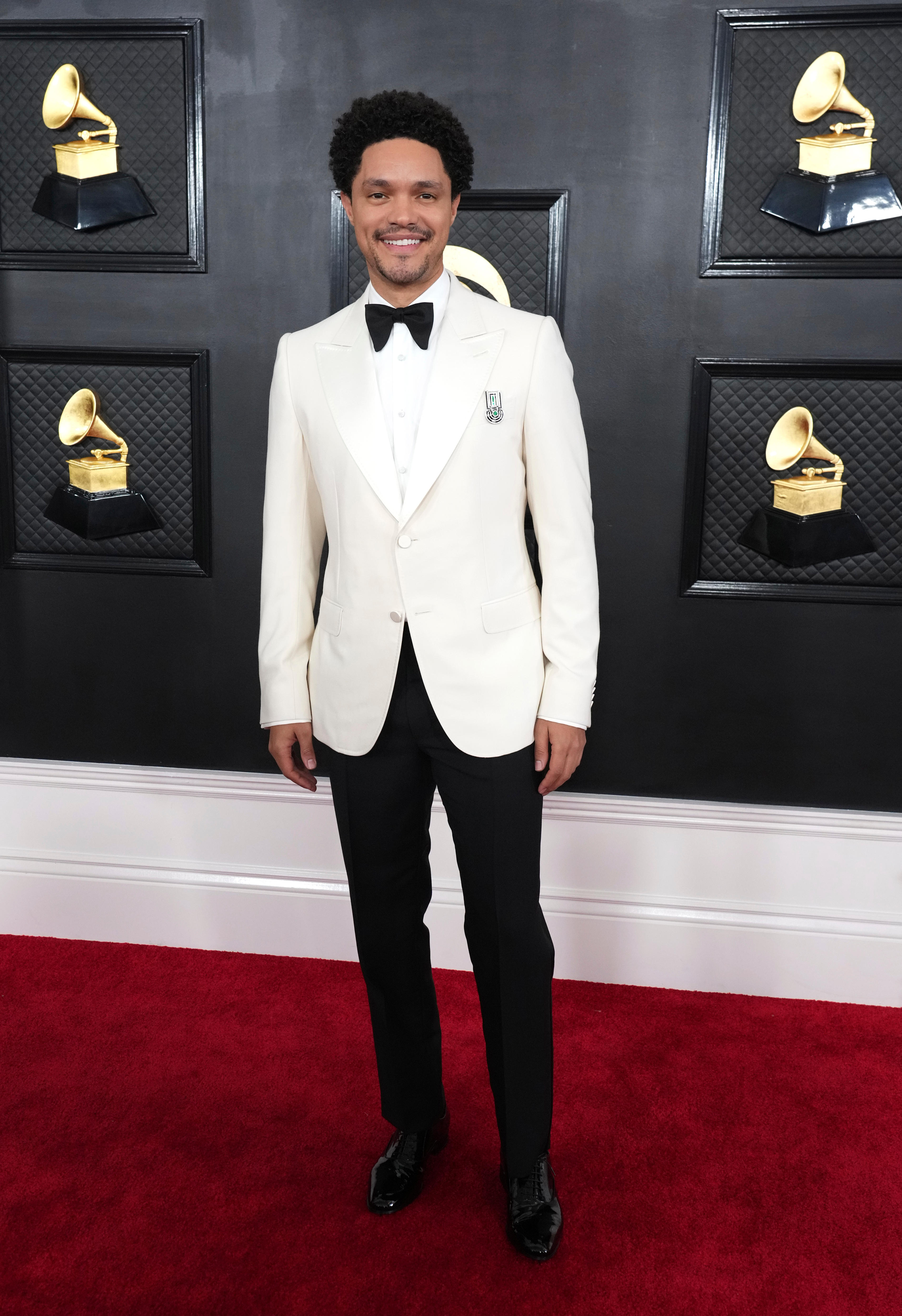 Trevor Noah wearing a white suit jacket and shirt with black trousers and an oversize black bow tie. 
