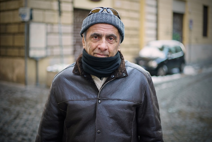 A middle-aged man wearing a beanie and scarf stands in an Italian street.