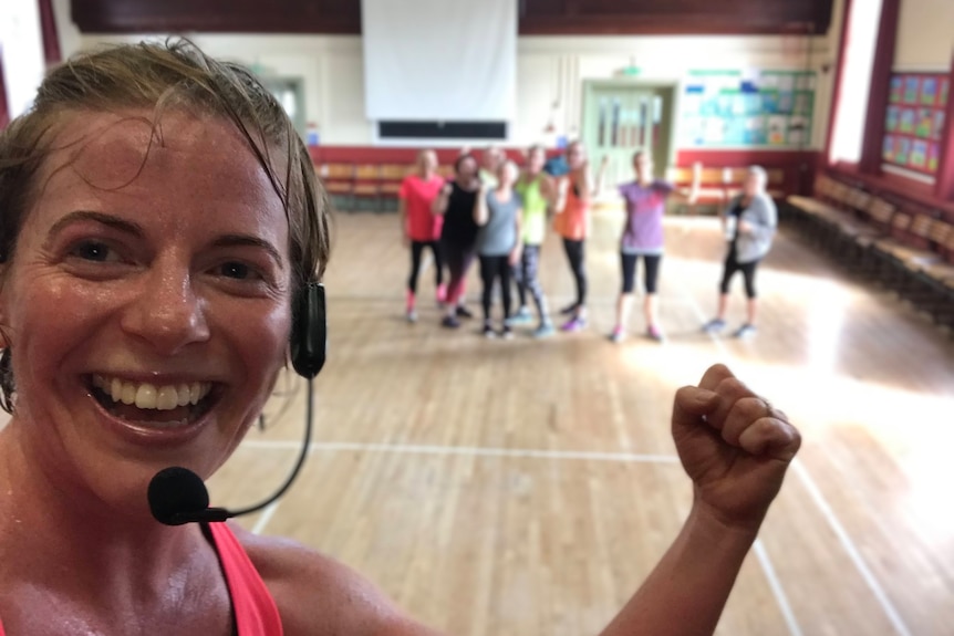 A group of women at an exercise class, one woman stands in focus n front, smiling to the camera. 