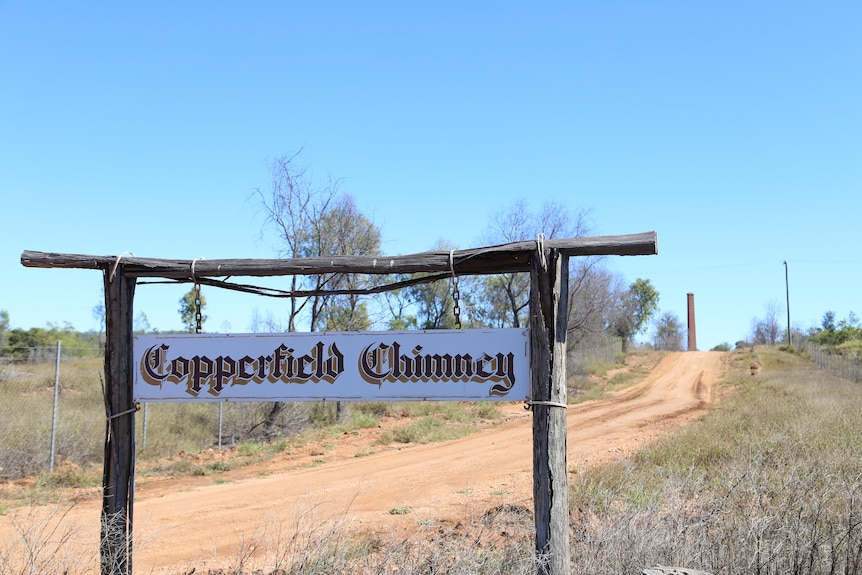 The words Copperfield Chimney hang from a wooden sign post at the start of a dirt road which leads to a red chimney stack.