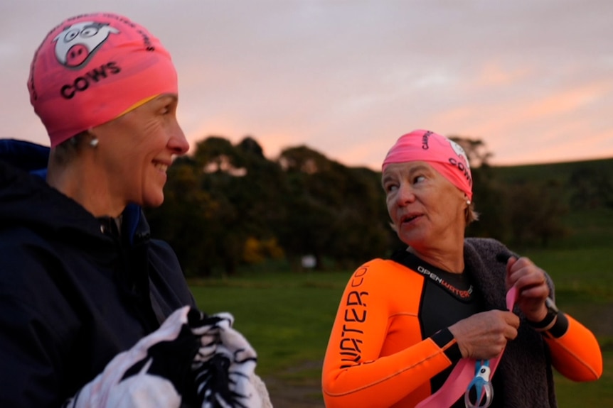 Two women, one in a wetsuit, wearing pink swimming caps that have a picture of a cow on them