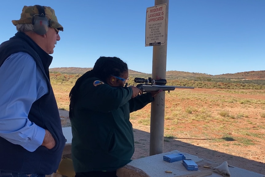 An Aboriginal women stands holding a gun to her shoulder aiming into the range. An man wearing earmuffs stands behind her.