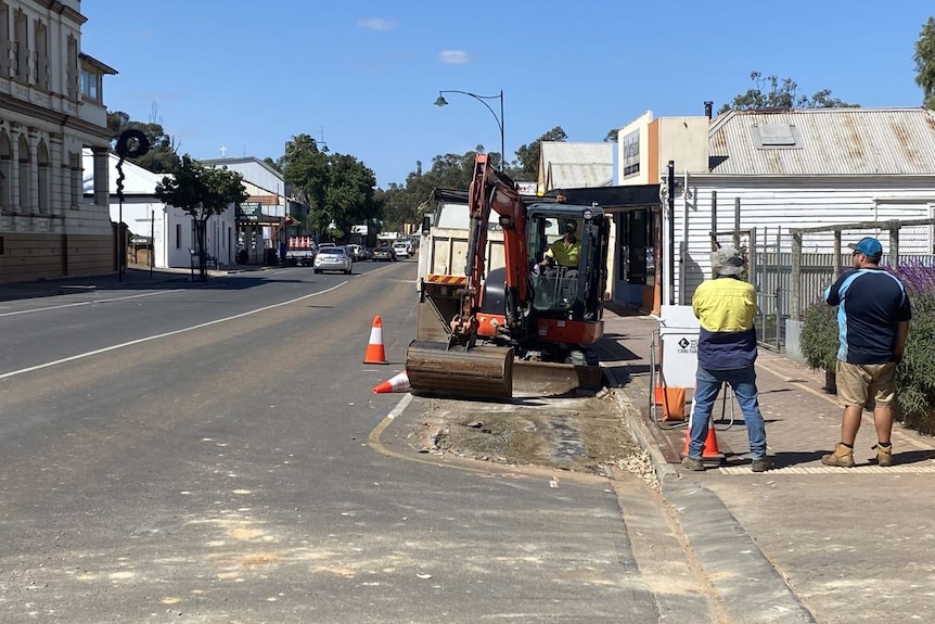 Two men stand looking at an excavator in a country town main street