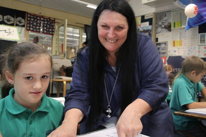 Yuluma Primary School Japanese teacher Margo Whittle kneels next to a student at her desk in the classroom.