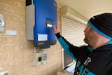 A man wears a black and teal beanie and jumper, he looks at a blue box mounted on a brick wall, he reads the small screen