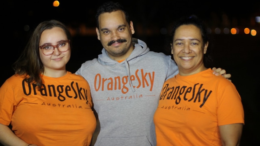 Rachael Musgrave stands smiling next to two other people. All are wearing Orange Sky t-shirts.