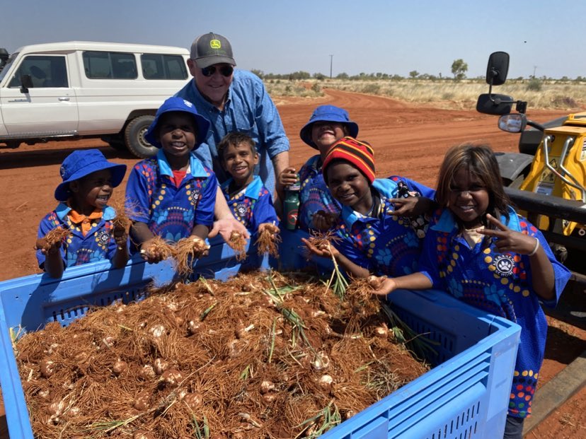 A man stands with children dressed in blue shirts and hats around a blue tub filled with dirt covered garlic bulbs.