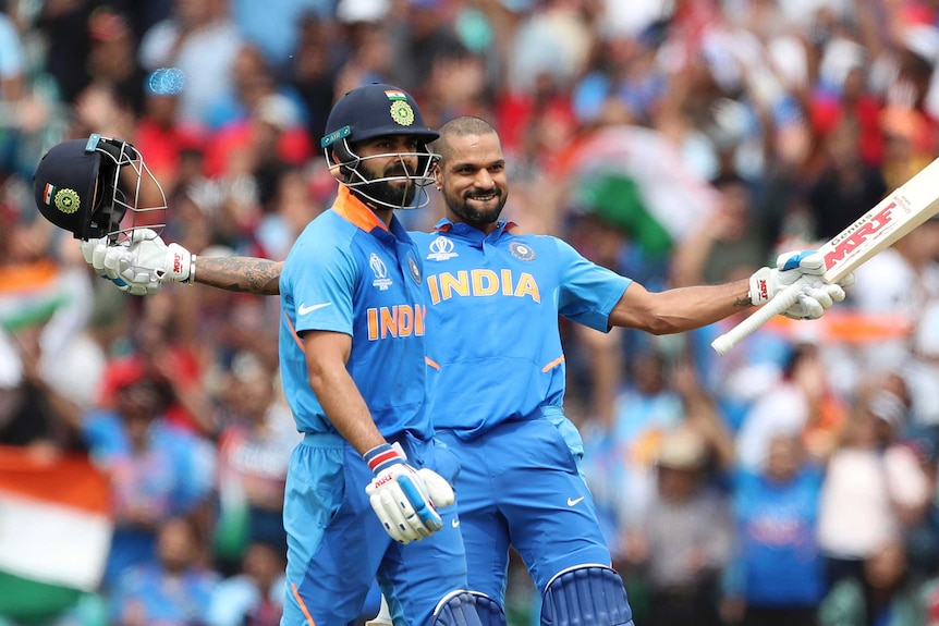 Shikhar Dhawan leans back with arms outstretched. He is smiling and has his helmet removed. Virat Kohli stands in the foreground