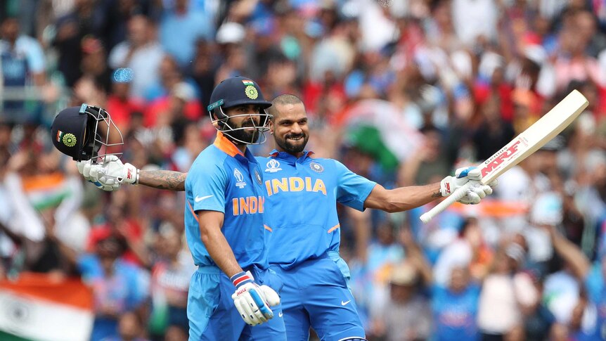 Shikhar Dhawan leans back with arms outstretched. He is smiling and has his helmet removed. Virat Kohli stands in the foreground