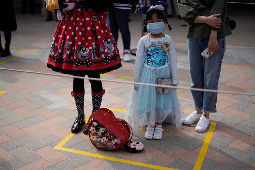 A small child wearing a Princess dress and a face mask stands in a yellow square at a theme park.