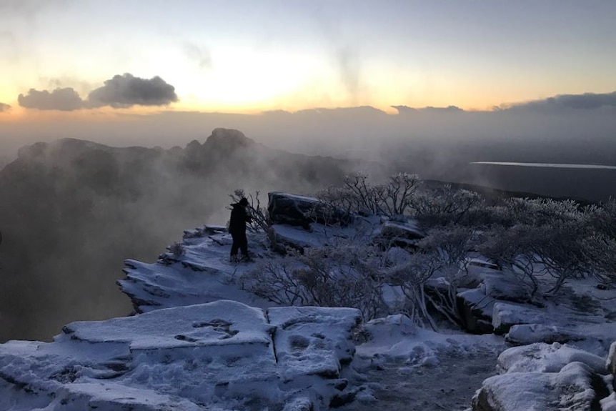 Light breaks revealing snow on the peak of Bluff Knoll and a man standing near the edge.