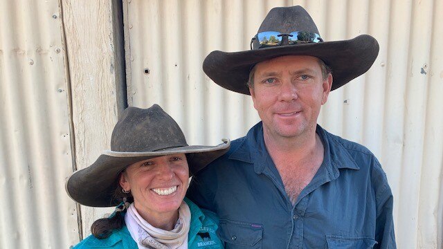 A man and woman wearing Akubras standing in front of corrugated iron, smiling