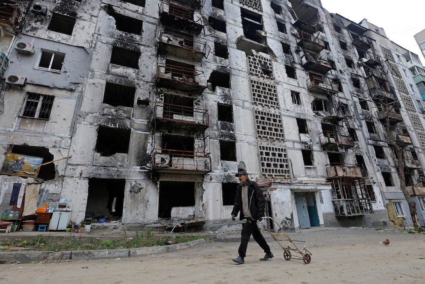 A man walks past a large damaged apartment building. 